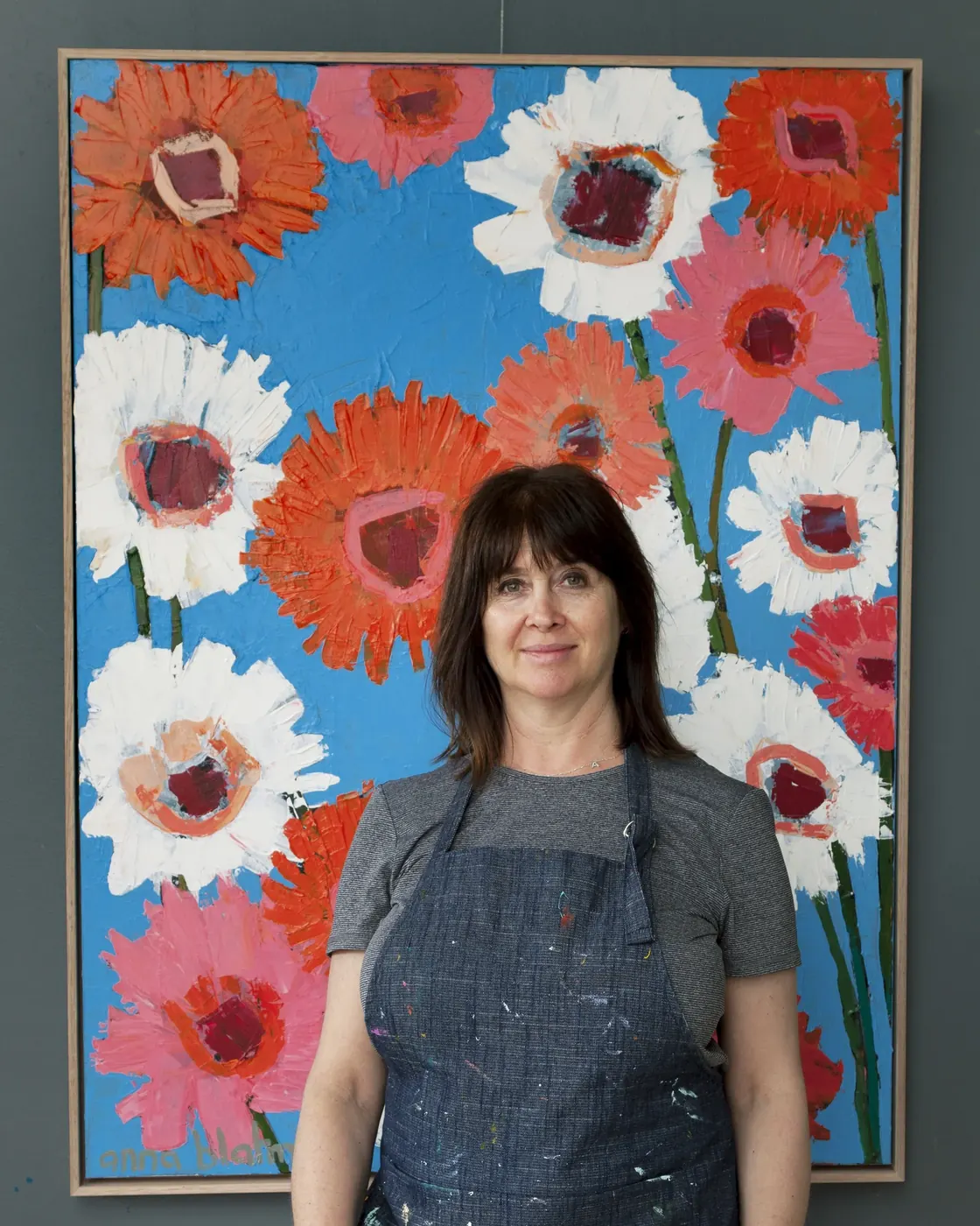 Anna Blatman standing in front of one of her large orange, pink and white gerbera paintings, wearing a paint-splattered denim apron