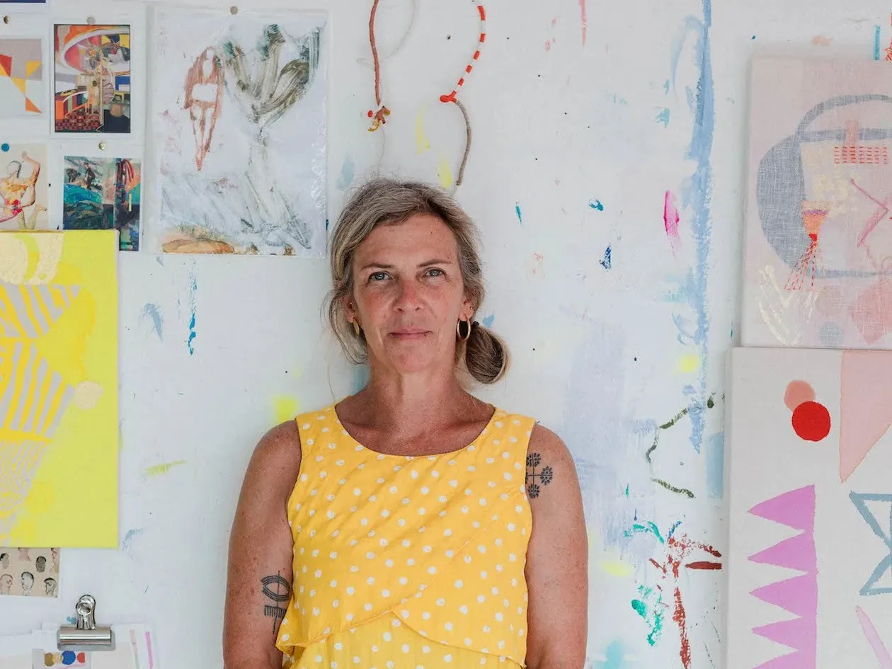 Becky Blair standing in a yellow polka dot dress in front of her paint-splattered Brighton studio wall, covered in colourful artworks and sketches