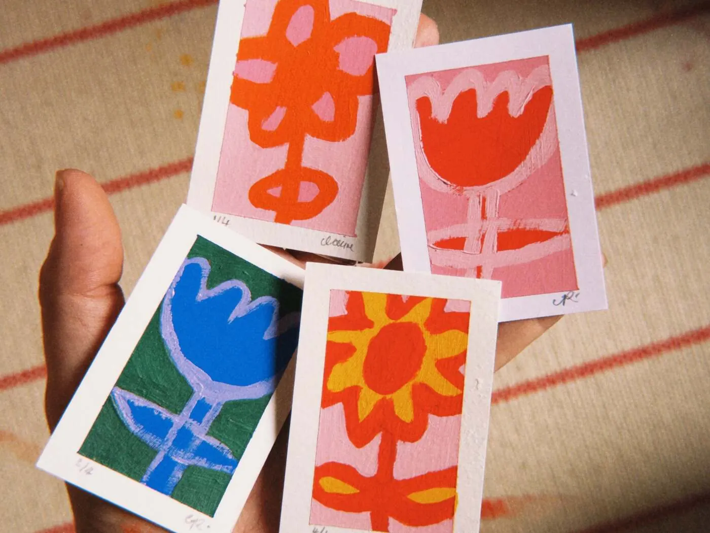 Four small hand-painted flower cards held in an open palm, in orange, pink, blue and green with scalloped petal edges