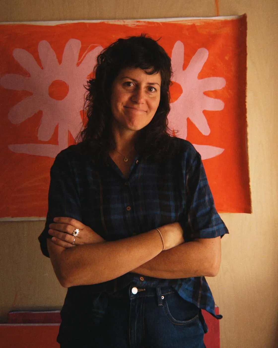 Claire Ritchie standing with arms crossed in front of a large orange painting with two pale pink daisy shapes, in her studio