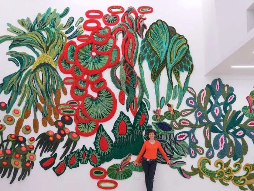 Claude Como standing in front of her tufted wool wall installation Sweet Symphony, with red, green and pink mushroom and plant shapes
