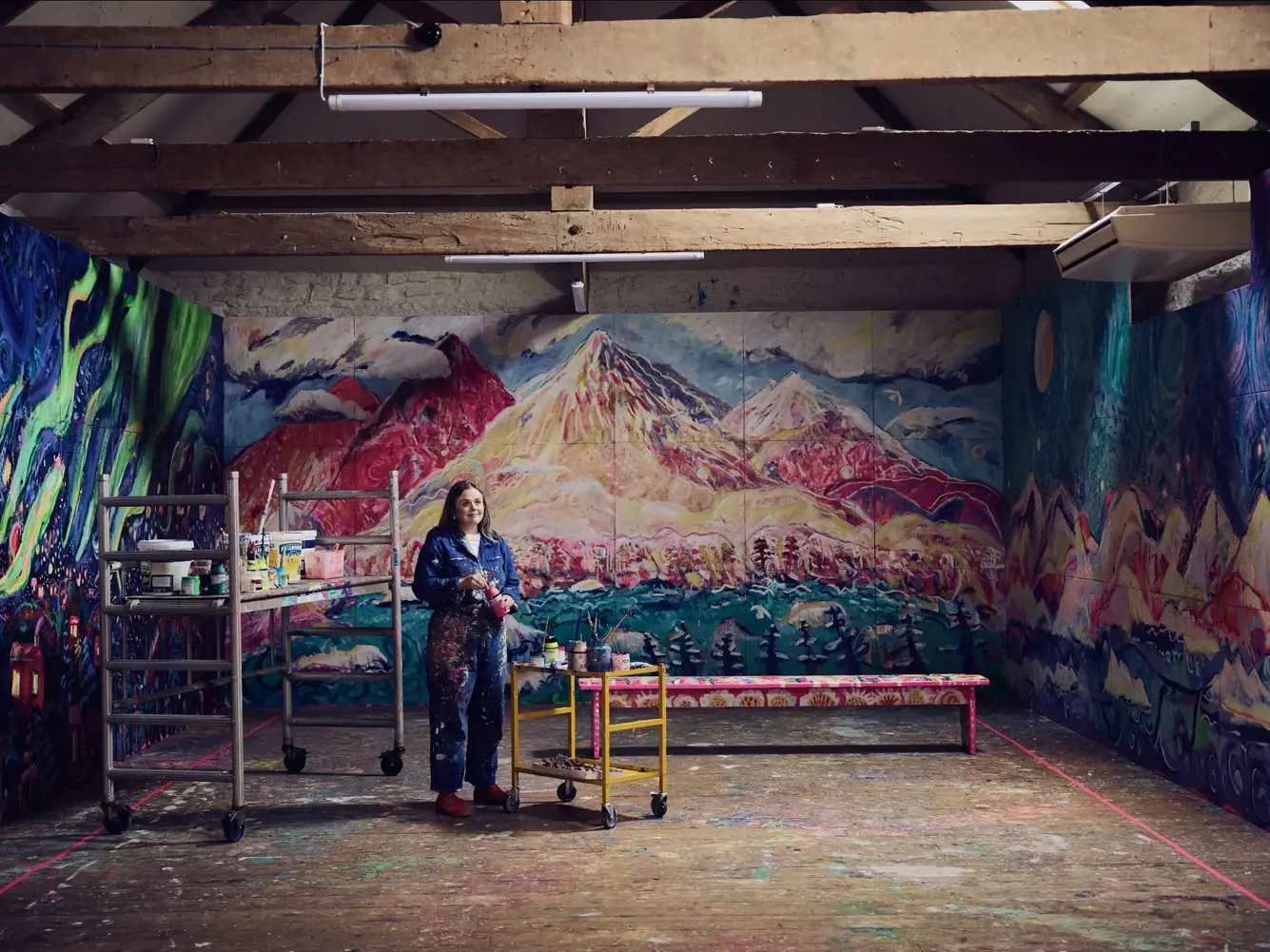 Emily Powell standing in her barn studio in Devon, surrounded by floor to ceiling paintings of pink and gold Arctic mountains over a teal forest