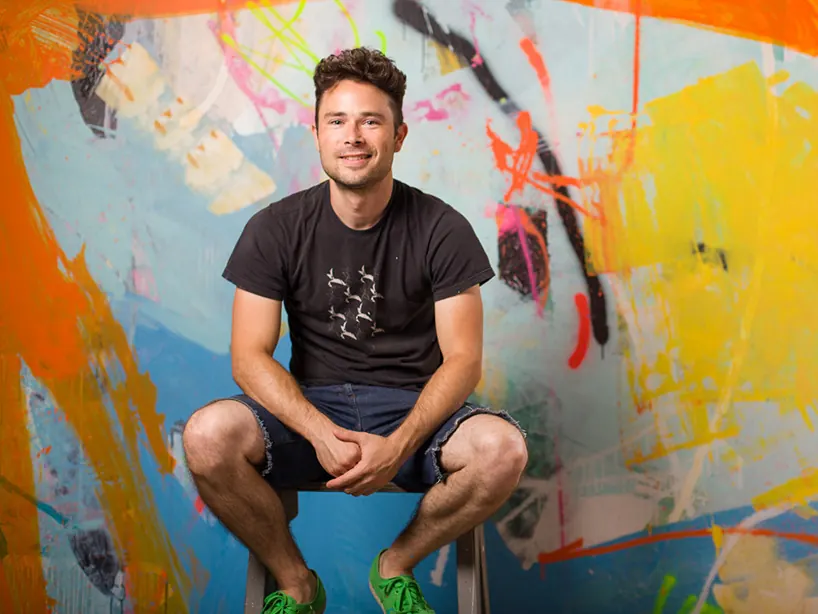 Alex Brewer, who paints as Hense, sitting on a stool in his Atlanta studio in front of a wall covered in orange, yellow and blue abstract brushwork
