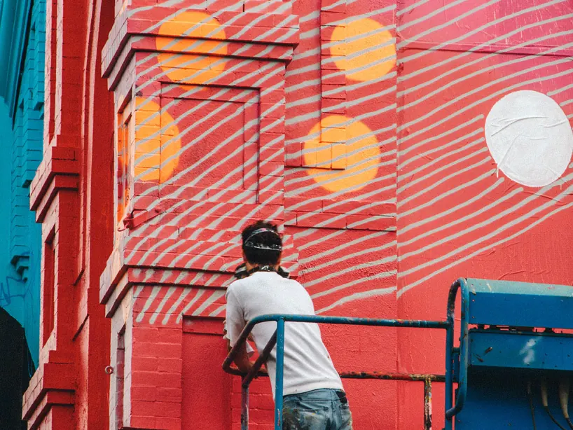Alex Brewer, painting as Hense, working from a lift on a red mural with orange circles and white spray-painted lines on a brick building