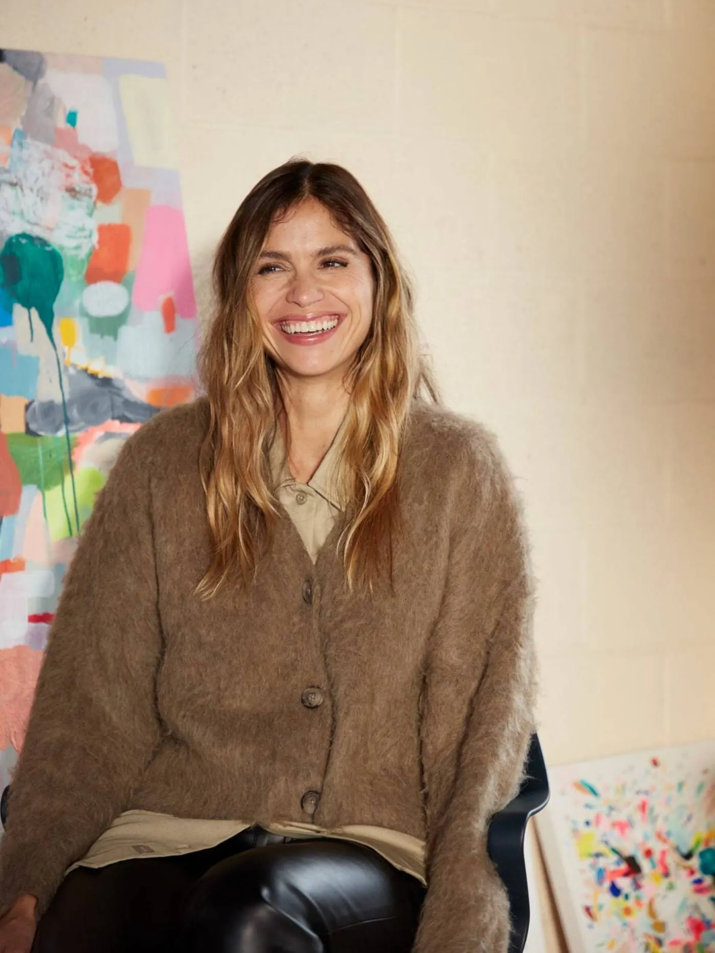 Kindah Khalidy smiling in her studio, with one of her paintings showing drips of pink, orange and green running down the canvas
