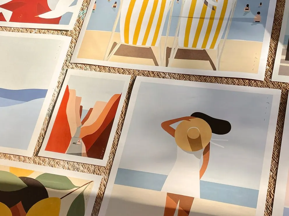 Prints by Quentin Monge laid out on a woven mat, showing striped beach chairs and a canyon road in flat colour