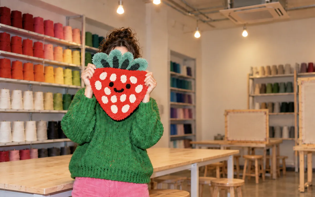A participant in the BUBU Studio holding up a tufted strawberry rug in front of the rainbow yarn wall